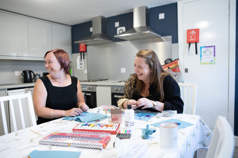 Two women sit at a kitchen table doing arts and crafts, They are laughing and looking to someone standing out of shot on the left.