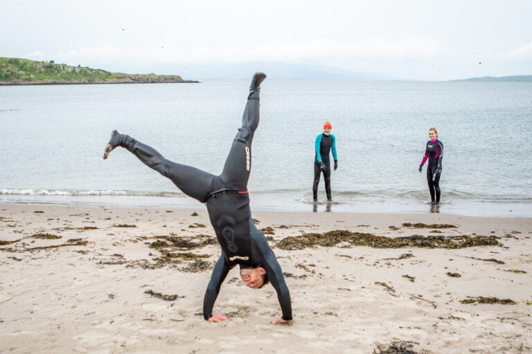 A young man does a handstand while wearing a wet suit on a beach. A man and a woman stand behind him, wearing wet suits in the sea.