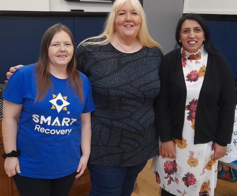 Three women stand together, smiling. One is wearing a SMART Recovery t-shirt.