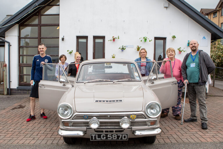 Six staff stand outside Oban Shore Street. There is a grey Ford Cortina four door saloon car. Three staff stand at one side, and three staff at the other side, with all the car doors open.