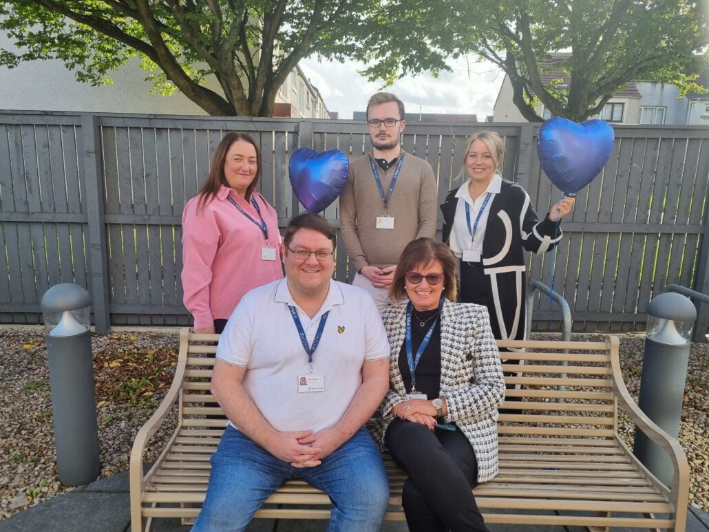 Five members of staff in a garden - two sit on a bench. Three people stand behind them, holding blue heart shaped balloons.
