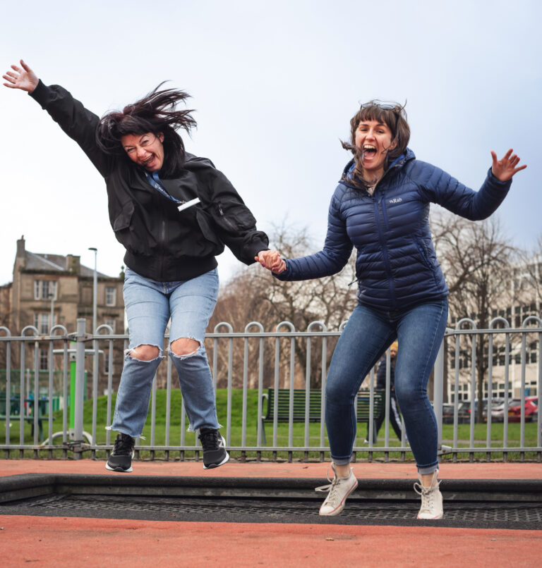 Two women jump in the air on a park trampoline.