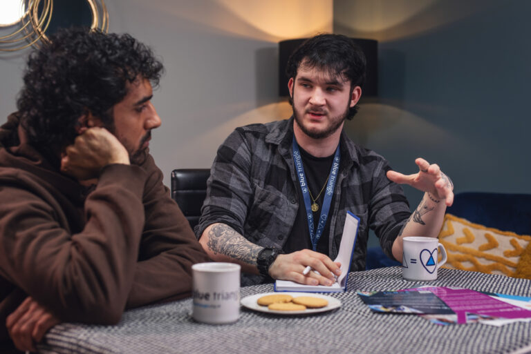 Blue Triangle staff member talks to supported person. They sit at a table with cups of tea and biscuits.