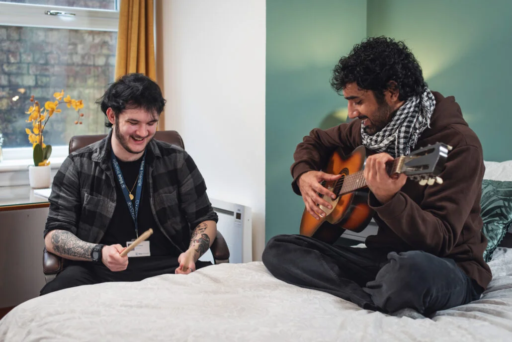 Young man sits on a bed with a guitar. A staff member sits beside him with a drumsticks.