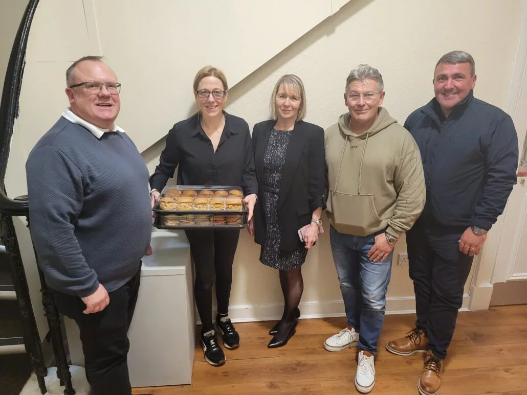 A group of people stand against stairs, one holding a platter of sandwiches.