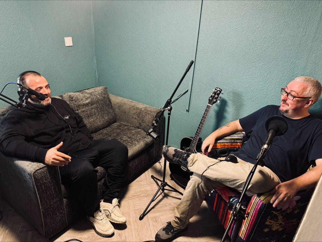 Image of two people talking in a studio sitting on a couch facing each other. Both have headphones on and speaking into a podcast microphone.