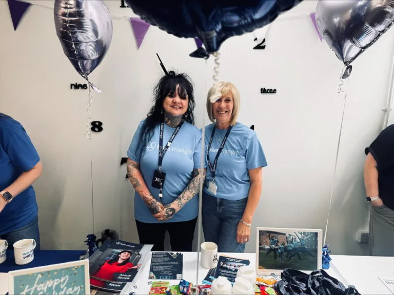 Two smiling people standing at a table with Blue Triangle tablecloth and branded goods, with balloons in the foreground.